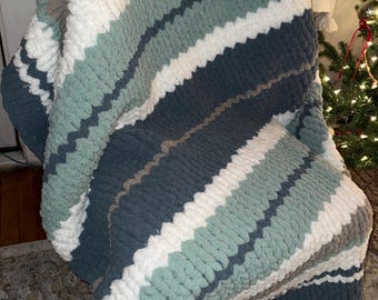 Puede incluir: Una manta de punto grueso a rayas en tonos azules, blancos y grises, colocada sobre una silla. La manta presenta amplias rayas de varios colores, creando una apariencia acogedora y texturizada. El material suave y afelpado sugiere calidez y confort.