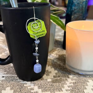 Puede incluir: Un soporte para bolsitas de té de metal plateado con una parte superior en espiral y una cadena colgante con pequeñas cuentas de piedra pulida de color azul claro. El soporte está unido a una etiqueta de té verde que dice "Steeped in Tradition Since 1945".