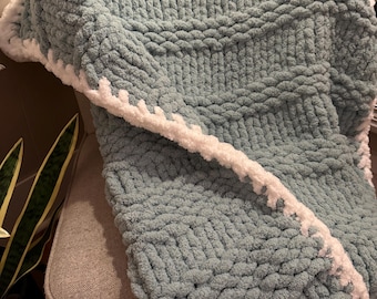 Puede incluir: Una manta de punto grueso en un suave color verde azulado con un borde blanco y esponjoso. La manta está sobre una silla, mostrando su tejido grueso y texturizado. Esta acogedora manta es perfecta para añadir calidez y estilo a cualquier espacio.