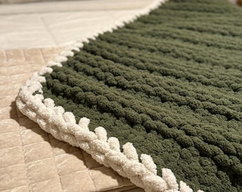 Puede incluir: Una manta tejida a mano en color verde oliva oscuro con un borde crema. La manta está hecha de hilo grueso y suave, perfecta para acurrucarse. Se muestra en una cama con una colcha y almohadas de tonos neutros.