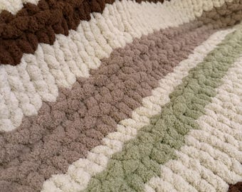 Puede incluir: Primer plano de una manta de punto grueso con rayas marrones, crema, verde claro y topo. La lana suave y texturizada crea una apariencia acogedora y acogedora. La manta es ideal para la decoración del hogar.