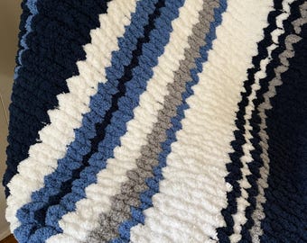 Puede incluir: Una manta de punto grueso con un diseño de rayas diagonales. La manta presenta rayas alternas en azul marino, azul claro, blanco y gris. La textura parece suave y acogedora, perfecta para acurrucarse.