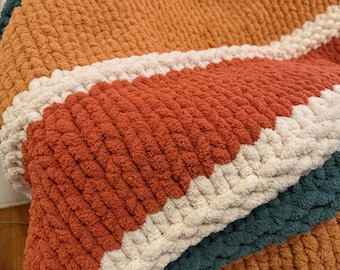 Puede incluir: Una manta tejida a mano con una textura gruesa. La manta presenta amplias rayas en tonos óxido, naranja, verde azulado y crema. El material suave y afelpado es perfecto para acurrucarse en el sofá o la cama.