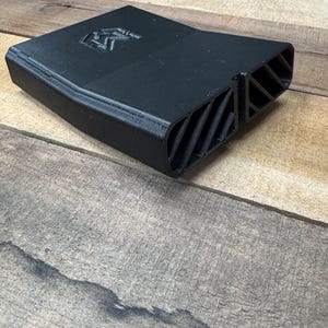 May include: A matte black 3D printed plastic component featuring a rectangular body with a tapered end. The top surface displays a debossed logo consisting of the letters RM. The front section features two distinct rectangular openings with diagonal slats.
