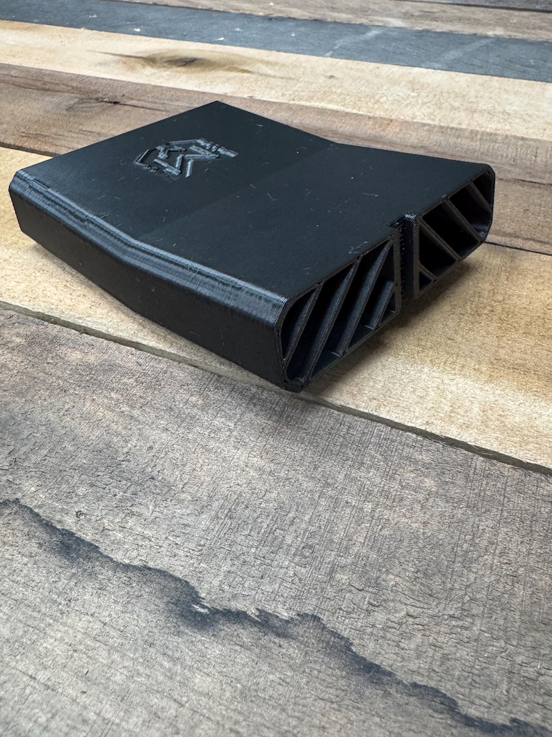 May include: A matte black 3D printed plastic component featuring a flat top surface with an embossed logo and a front section with two rectangular openings containing diagonal slats. The object has a tapered, angular shape and a textured finish.
