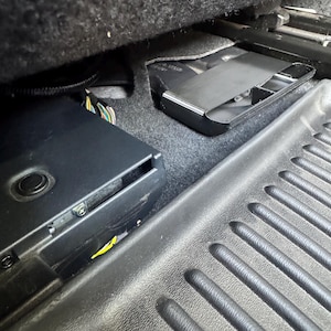 May include: A black electronic control module with a circular button and visible metal screws, positioned under a vehicle seat next to a black plastic floor mat with a ribbed texture and a secondary black plastic component nearby.