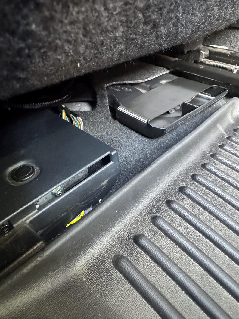 May include: A black electronic control module with a circular button and visible metal screws, positioned under a vehicle seat next to a black plastic floor mat with a ribbed texture and a secondary black plastic component nearby.