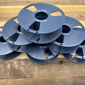 May include: A stack of six dark blue plastic 3D printer filament spools. Each circular spool features a perforated design with a central hub and multiple cutouts along the outer rim, arranged in a pyramid formation on a wooden surface.