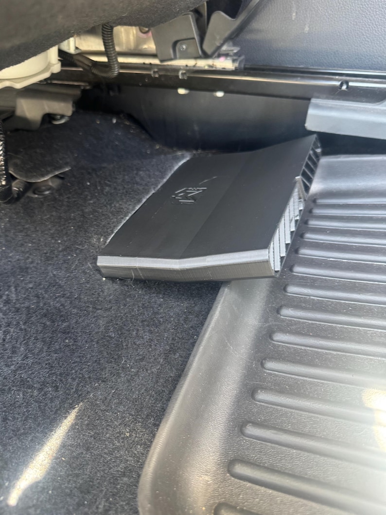 May include: A black plastic automotive air vent cover with a geometric design and a visible logo, positioned on the dark carpeted floor of a vehicle interior next to a black rubber floor mat with raised horizontal ridges and a smooth border.