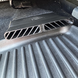 May include: A black plastic automotive air vent extension featuring a series of diagonal slats and a central divider. The component is designed with a rectangular opening and a textured surface, positioned directly over a ribbed black floor mat.