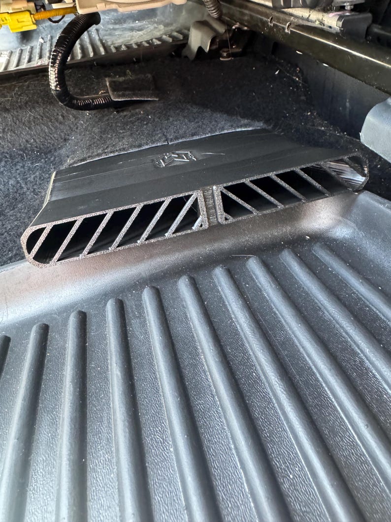 May include: A black plastic automotive air vent extension featuring a series of diagonal slats and a central divider. The component is designed with a rectangular opening and a textured surface, positioned directly over a ribbed black floor mat.
