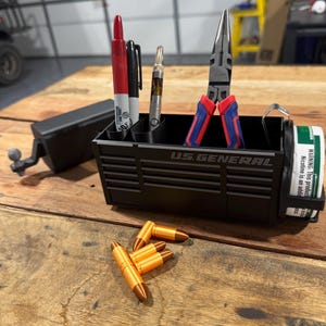 May include: Black organizer with the text "U.S. GENERAL" holding pens, a vape pen, and pliers. Several orange and bronze-colored bullet-shaped objects are on the wooden surface. A black box with a crank is on the left.