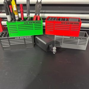 May include: A collection of four miniature tool chest organizers in green, red, black, and gray, each featuring the text U.S. GENERAL on the front. These rectangular containers have multiple compartments for storing various hand tools and pens.