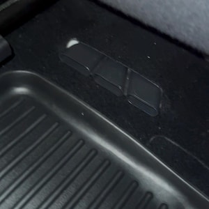 May include: A black automotive floor mat with a ribbed texture positioned on a dark carpeted surface. The mat features three distinct, rectangular, hollow plastic air vent openings integrated into the floor area near the edge of the mat.