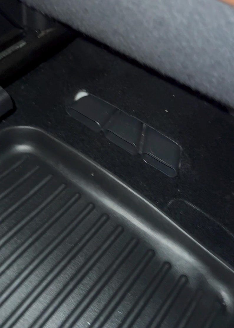 May include: A black automotive floor mat with a ribbed texture positioned on a dark carpeted surface. The mat features three distinct, rectangular, hollow plastic air vent openings integrated into the floor area near the edge of the mat.