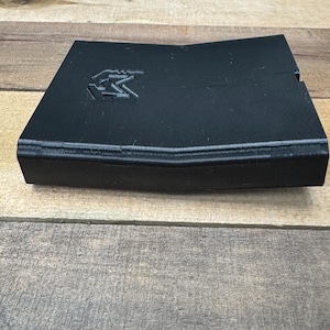 May include: A black, angular, 3D-printed plastic object with a slightly bent, V-shaped profile. The top surface features a debossed logo consisting of the letter M inside a shield-like outline. The object has a matte finish and visible layer lines.