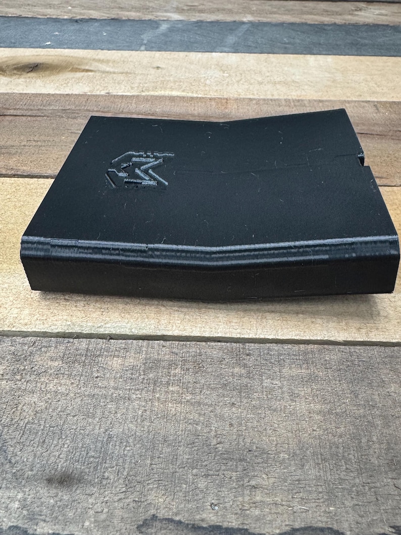 May include: A black, angular, 3D-printed plastic object with a slightly bent, V-shaped profile. The top surface features a debossed logo consisting of the letter M inside a shield-like outline. The object has a matte finish and visible layer lines.