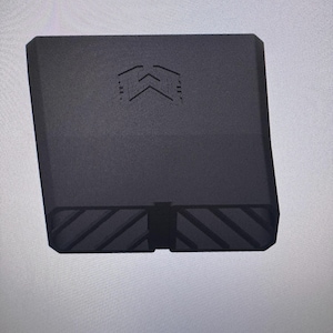 May include: A matte black, rectangular, hollow component featuring a geometric logo on the front face. The bottom edge displays a series of diagonal, recessed grooves across its width, creating a textured pattern on the base of the object.