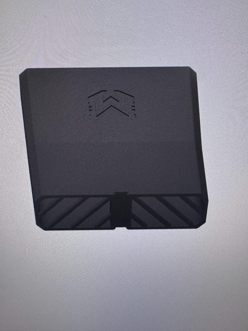 May include: A matte black, rectangular, hollow component featuring a geometric logo on the front face. The bottom edge displays a series of diagonal, recessed grooves across its width, creating a textured pattern on the base of the object.
