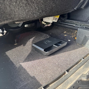 May include: A black plastic storage tray with a rectangular shape and a molded logo on the top surface, positioned on the dark gray carpeted floor of a vehicle interior, located directly underneath the front seat assembly and wiring components.