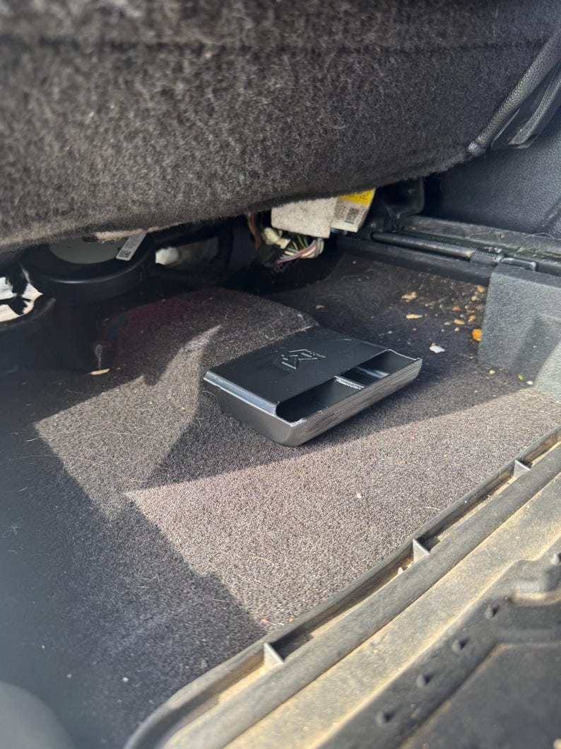 May include: A black plastic storage tray with a rectangular shape and a molded logo on the top surface, positioned on the dark gray carpeted floor of a vehicle interior, located directly underneath the front seat assembly and wiring components.