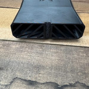 May include: A matte black 3D printed rectangular duct component featuring a central vertical divider and two side openings with internal diagonal support ribs. The top surface displays a stamped letter M logo centered near the edge.