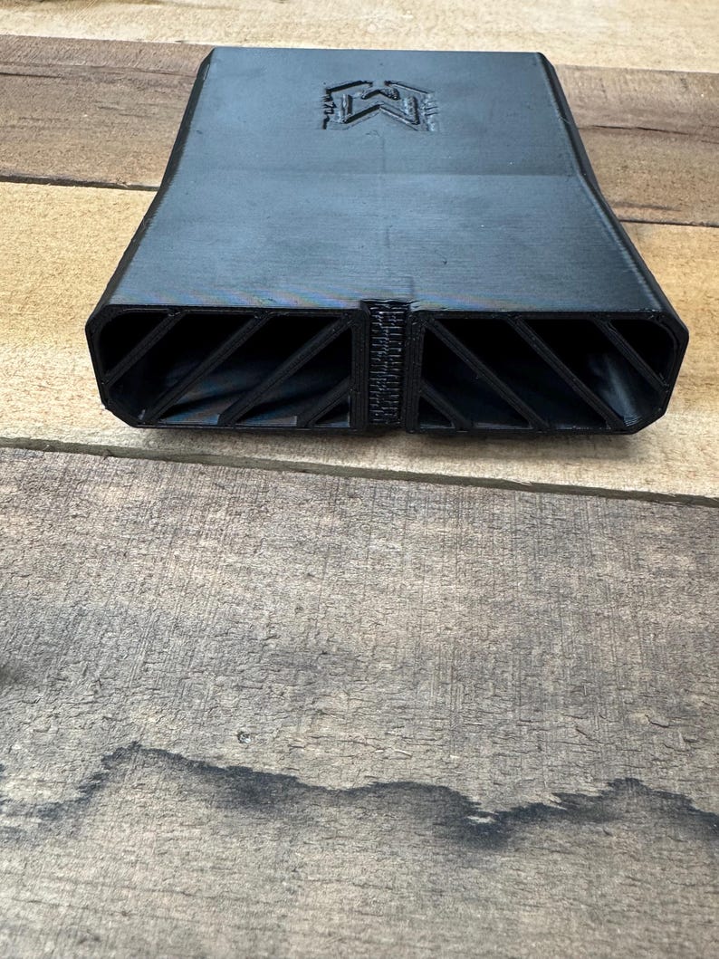 May include: A matte black 3D printed rectangular duct component featuring a central vertical divider and two side openings with internal diagonal support ribs. The top surface displays a stamped letter M logo centered near the edge.
