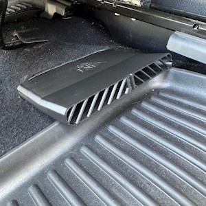 May include: A black plastic car air vent cover with a slatted design and a small logo embossed on the top surface. The rectangular accessory is positioned on a black rubber floor mat beneath a vehicle seat, featuring a series of angled vents.