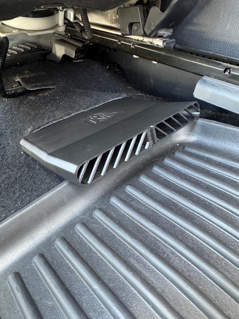 May include: A black plastic car air vent cover with a slatted design and a small logo embossed on the top surface. The rectangular accessory is positioned on a black rubber floor mat beneath a vehicle seat, featuring a series of angled vents.