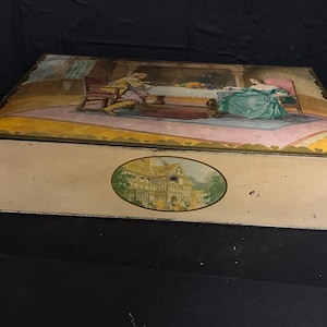 May include: Vintage rectangular tin box with a cream-colored base and a decorative lid. The lid features a painting of a dining scene with figures and a landscape scene on the front. The box has a gold-colored trim.