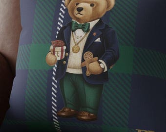 May include: Decorative pillow featuring a teddy bear wearing a navy blazer, green pants, and a bow tie. The bear is holding a gift and a gingerbread man. The pillow has a plaid pattern in navy, green, and white.