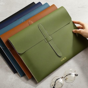 Peut inclure: Plusieurs pochettes en cuir de différentes couleurs : noir, bleu marine, turquoise, marron et vert olive. Le porte-documents vert olive porte les initiales "JJKG" en or. Des lunettes rondes sont posées à côté.