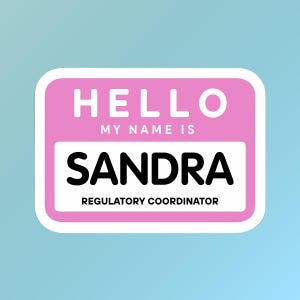 May include: A pink and white name tag sticker with rounded corners. The text "HELLO" is at the top, followed by "MY NAME IS" and the name "SANDRA" in bold black letters. Below, it reads "REGULATORY COORDINATOR."