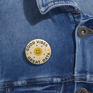 Könnte beinhalten: Ein runder, cremefarbener Button mit den Worten "GOOD VIBES GREAT DATA", die ein lächelndes Sonnensymbol umgeben. Der Button ist mit kleinen blauen und gelben Punkten verziert. Der Button ist an einer blauen Jeansjacke befestigt.