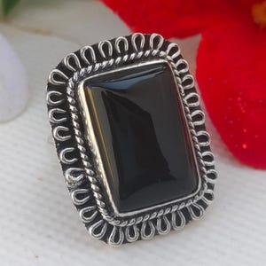 Könnte beinhalten: Rechteckiger Ring aus schwarzem Onyx mit einer silberfarbenen, verzierten Fassung. Der Ring hat einen großen, polierten schwarzen Stein, der in einen dekorativen Rahmen mit einem seilartigen Rand und einem gewellten Rand gefasst ist.