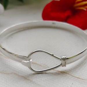 May include: A silver bangle bracelet with a hook and eye clasp. The bracelet has a smooth, polished finish. The clasp is a simple, elegant design. The background includes a red flower. The bracelet is a classic piece of jewellery.