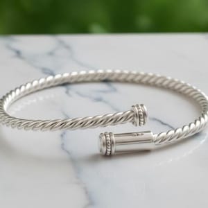 Könnte beinhalten: Ein silberfarbenes, offenes Armreif-Armband mit einem gedrehten Seil-Design. Das Armband hat einen Verschluss und wird auf einer weißen Marmoroberfläche präsentiert. Es ist ein Schmuckstück.