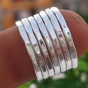 Puede incluir: Primer plano de una pila de anillos de plata. Los anillos son lisos y brillantes, con una textura martillada. Los anillos están apilados en un dedo, con un fondo verde borroso.