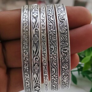 Pode incluir: Um conjunto de seis pulseiras de prata com intrincados padrões florais e de folhas. As pulseiras são seguradas em uma mão, mostrando as gravuras detalhadas e o acabamento polido da joia. O design inclui flores, vinhas e outros elementos decorativos.
