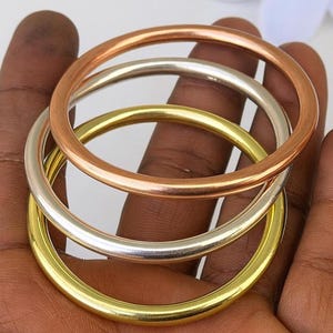 Könnte beinhalten: Drei Metallarmreifen in Kupfer-, Silber- und Goldtönen. Die Armreifen sind rund und glatt, mit einem schlichten Design. Der kupferfarbene Armreif liegt oben, gefolgt von einem silbernen und dann einem goldenen Armreif. Die Armreifen sind gestapelt.