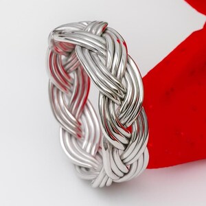 May include: A silver ring featuring a woven or braided design. The ring's surface is textured, created by multiple strands intertwined. The background is white and red, highlighting the ring's metallic sheen.