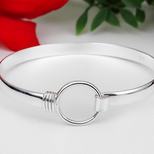 May include: A silver bangle bracelet with a circular clasp. The bracelet is made of polished metal and features a simple, elegant design. The clasp is a prominent feature, adding a modern touch to the classic style. The bracelet is shown against a soft, neutral background.
