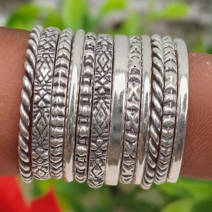 May include: A collection of silver rings with various textures and patterns. The rings include twisted rope, geometric designs, and smooth bands. The rings are worn on a finger, showcasing the different styles and textures of the jewelry.