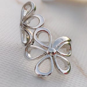 May include: A silver-colored floral ring featuring two openwork flower designs. The ring has a polished finish and is set against a soft, white background. The flowers have rounded petals and a central detail, creating a delicate and elegant look.