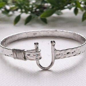 May include: A silver-colored bracelet with a hammered texture and a hook clasp. The bracelet has a rounded shape and a textured surface. The hook clasp is a U-shape. The bracelet is a piece of jewelry.