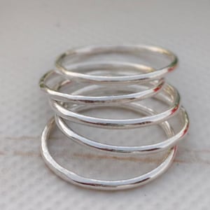 Pu&ograve; includere: Una pila di cinque anelli d'argento. Ogni anello presenta una texture martellata e un design minimalista. Gli anelli sono impilati, creando un effetto a strati. Sfondo chiaro.