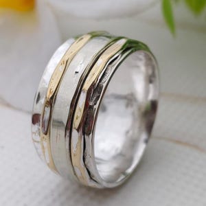 May include: A silver ring with gold bands. The ring has a textured surface and a wide band design. The gold bands are placed between the silver bands. The ring is a piece of jewellery.