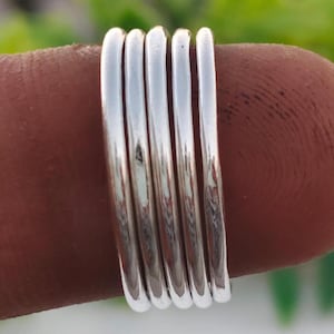 Pu&ograve; includere: Una pila di cinque anelli d'argento, ciascuno con un design liscio e arrotondato. Gli anelli sono mostrati su un dito, con uno sfondo verde sfocato. Gli anelli sembrano essere fatti di metallo lucido.