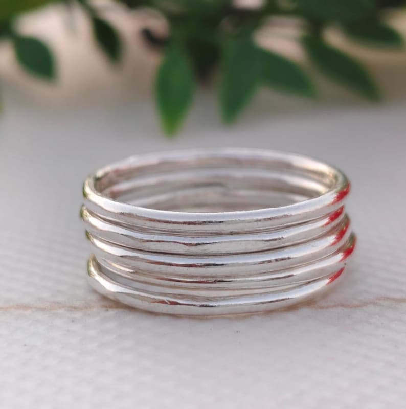 Pu&ograve; includere: Una pila di sei anelli d'argento. Gli anelli hanno una texture martellata e una finitura lucida. Gli anelli sono impilati su uno sfondo sfocato con fogliame verde. Ogni anello misura circa 0,2 cm di spessore.