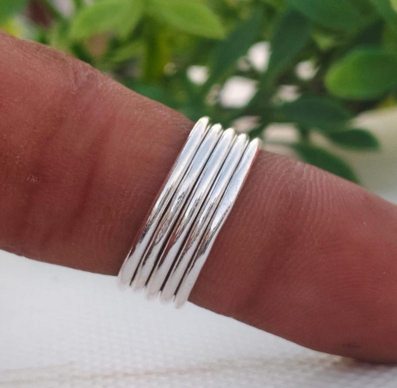 Pu&ograve; includere: Una pila di sei anelli color argento, ciascuno con una superficie liscia e lucida. Gli anelli sono indossati su un dito, con uno sfondo verde sfocato. Gli anelli sono semplici ed eleganti, adatti all'uso quotidiano.